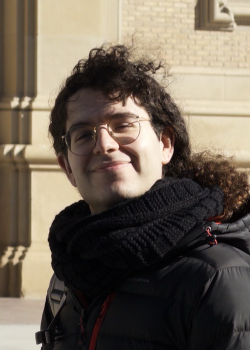 The founder of Onanem City, circa 2026. A cute boy with long curly dark brown hair stands in front of the Cathedral-Basilica of Our Lady of the Pillar in Zaragoza. He wears a black knitted scarf and a black puffer jacket. He smiles to camera while the wind waves his curls and the sun shines on the side of his face, for a short lighting feel.